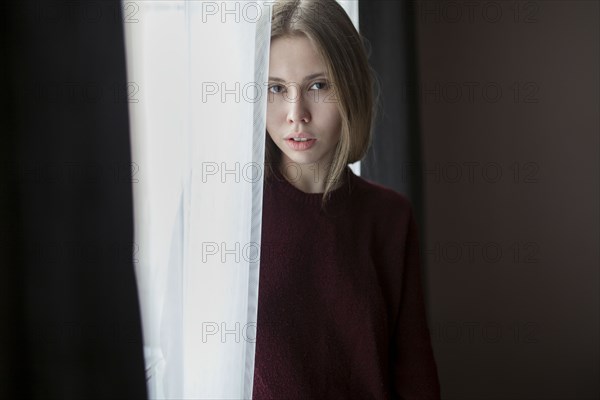 Serious Caucasian woman standing near curtain at window