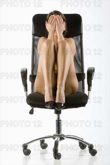 Naked Caucasian woman wearing high heels covering face in chair