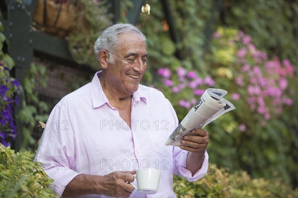 Hispanic man drinking coffee and reading newspaper in flower garden