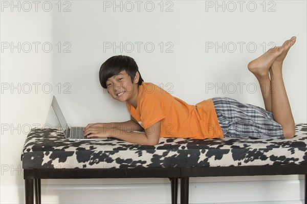 Smiling Asian boy laying on bench using laptop
