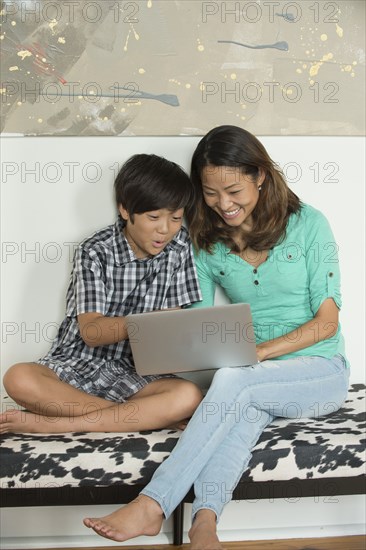 Smiling Asian mother and son sitting on bench using laptop