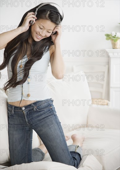 Asian woman wearing headphones and kneeling on sofa