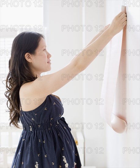 Pregnant Asian woman decorating room with wallpaper