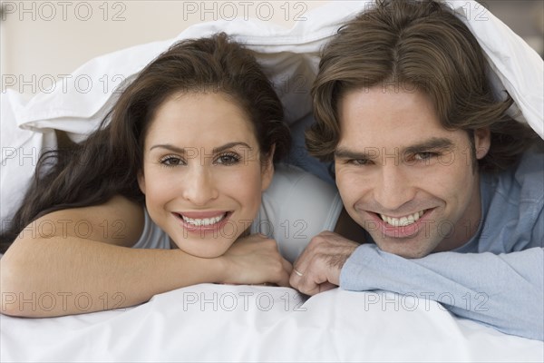 Hispanic couple smiling from under covers on bed