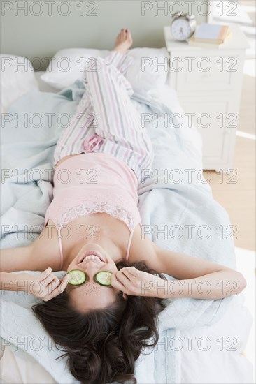 Mixed race woman placing cucumbers over eyes in bed