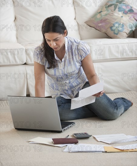 Hispanic woman paying bills with laptop on living room floor