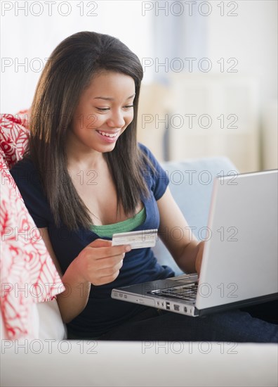 Smiling mixed race teenage girl holding credit card and laptop on sofa