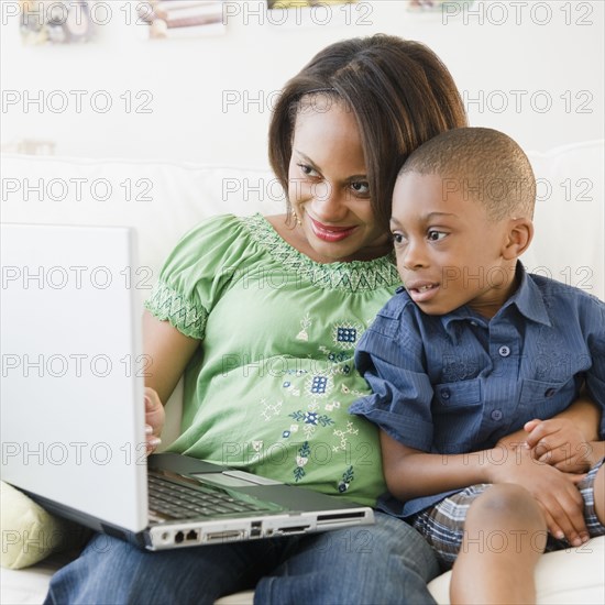 Black mother and son using laptop on sofa