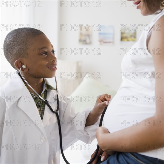 Black boy in lab coat holding stethoscope to pregnant mother's stomach