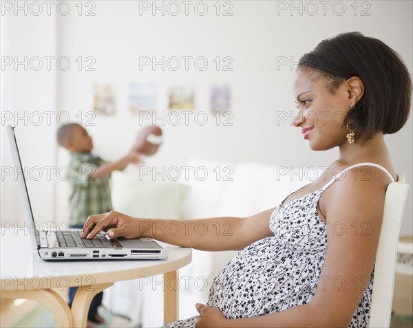 Pregnant Black mother using laptop while son plays with football