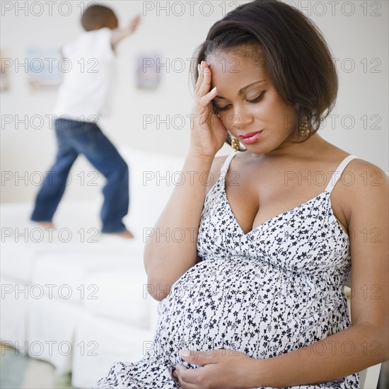 Pregnant Black mother sitting with head in hands while son jumps on bed