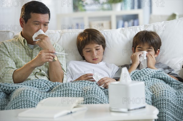 Sick father and sons blowing noses and checking temperature on living room sofa