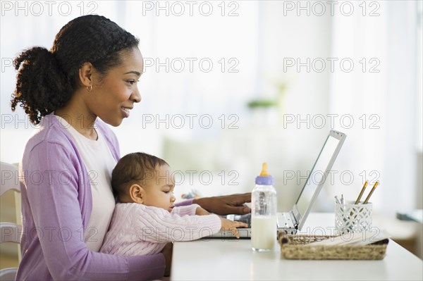 Mixed race woman holding baby and using laptop