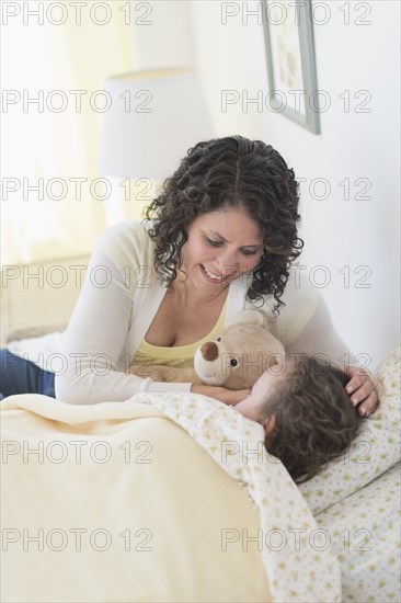 Mixed race mother tucking daughter and teddy bear into bed