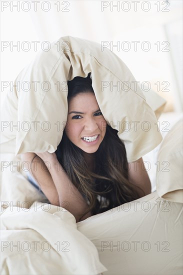 Portrait of frustrated Hispanic woman under pillow in bed