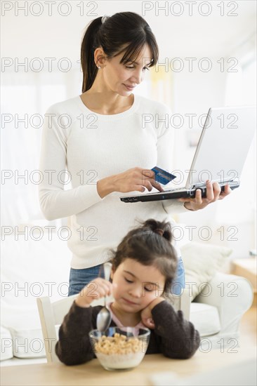 Hispanic mother shopping online while daughter eats cereal