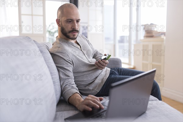 Hispanic man using cell phone and laptop on sofa