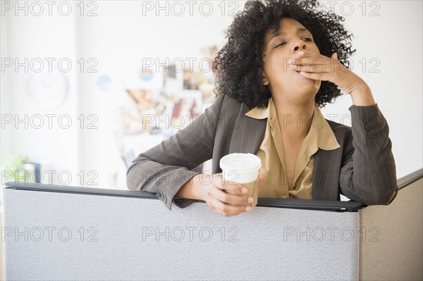 Businesswoman with cup of coffee yawning in cubicle