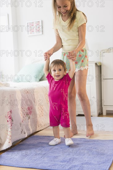 Caucasian girl helping toddler sister walk in bedroom