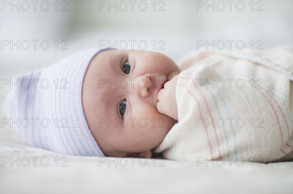 Swaddled Asian baby biting fingers on bed