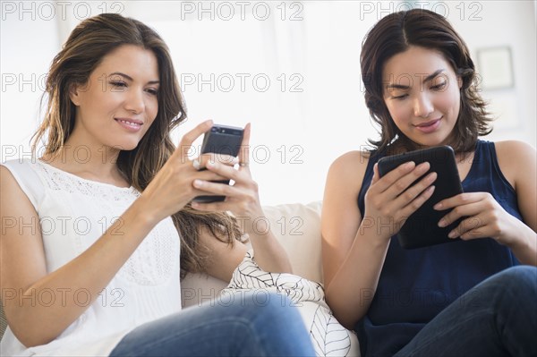 Hispanic women using cell phone and digital tablet on sofa