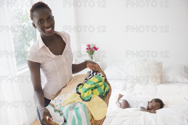 Black mother carrying laundry near sleeping baby boy in bedroom