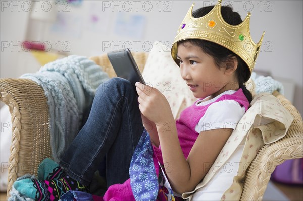 Vietnamese girl with crown using digital tablet in armchair