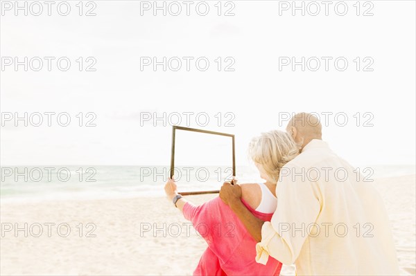 Older couple holding empty picture frame on beach