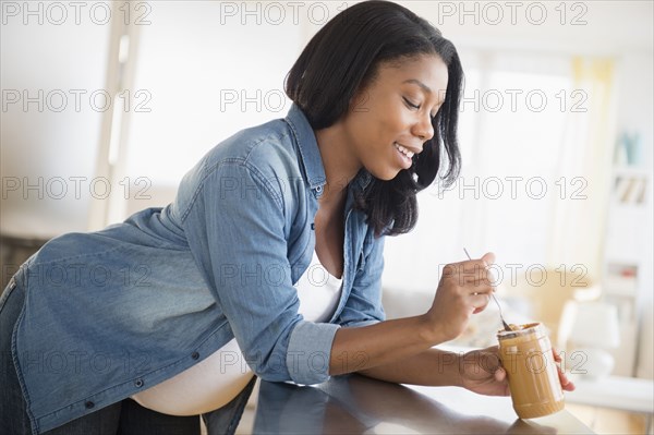 Black pregnant woman eating peanut butter in kitchen