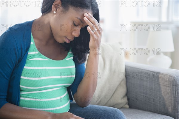 Anxious Black pregnant woman rubbing forehead on sofa