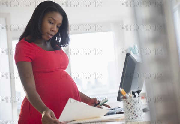 Black pregnant businesswoman reading paperwork at desk in office