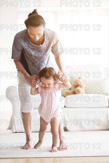 Mother helping baby daughter walk in living room