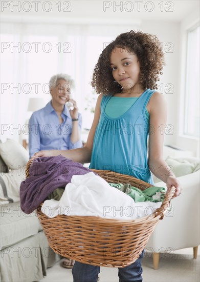 Girl holding laundry basket with grandmother on phone in background