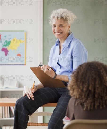 Mixed race teacher holding clipboard and smiling at student in classroom
