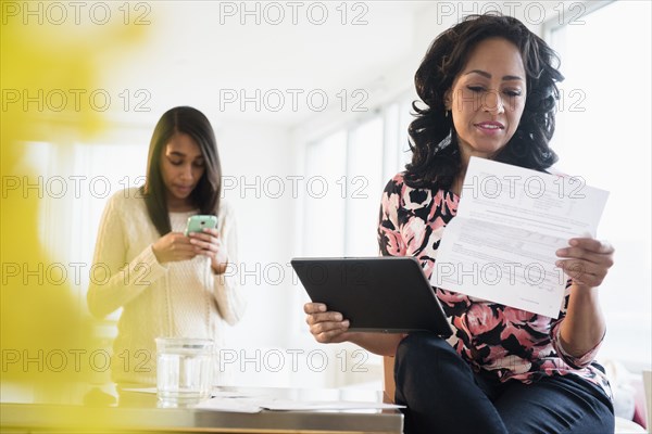 Mother paying bills with digital tablet while daughter uses cell phone