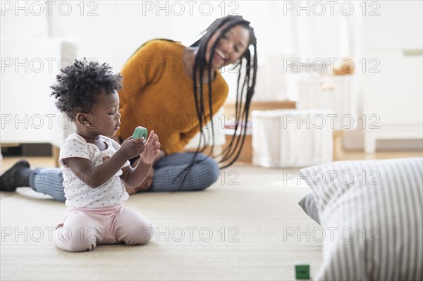 Black woman watching baby daughter play with toy on carpet