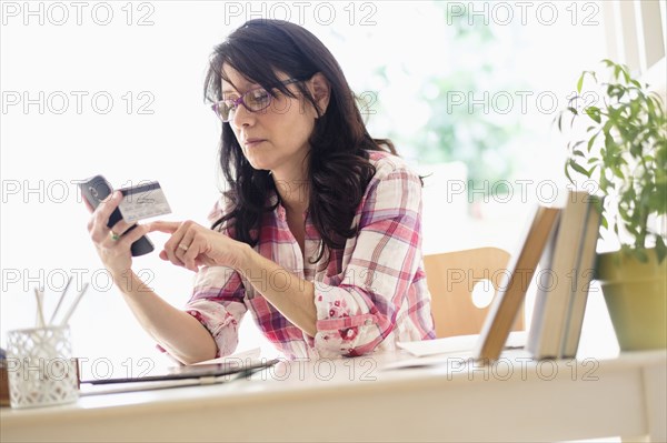 Hispanic woman using credit card with cell phone in home office