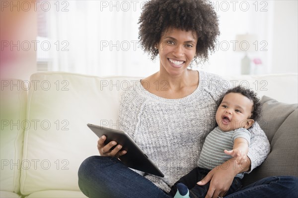 Mother sitting on sofa holding baby son and digital tablet