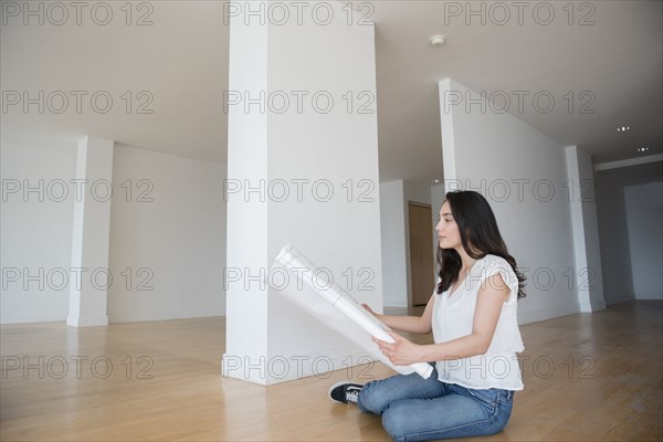 Hispanic woman sitting on floor of empty apartment holding blueprint