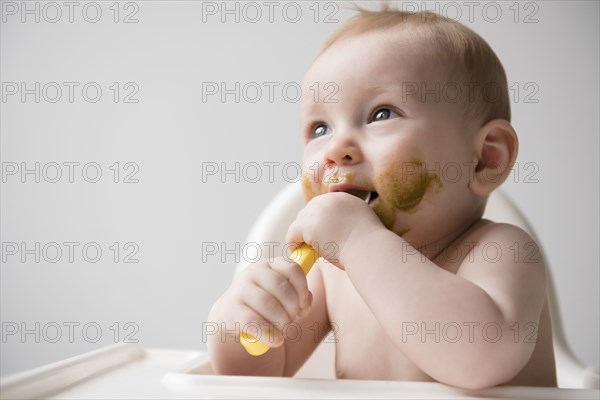 Messy Caucasian baby boy eating from spoon in high chair