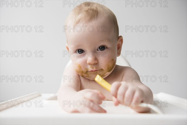 Messy Caucasian baby boy holding spoon in high chair