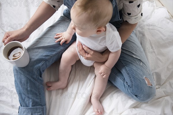 Caucasian mother holding baby son in lap on bed while drinking coffee