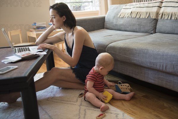 Caucasian mother sitting on floor using laptop near baby son