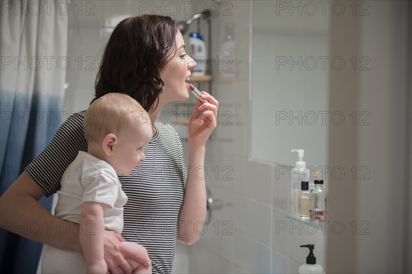 Caucasian mother holding baby son while applying lipstick in mirror