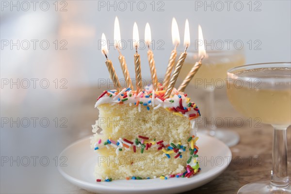 Candles burning on slice of cake with sprinkles near champagne