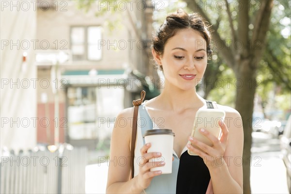 Thai woman holding coffee texting on cell phone in city