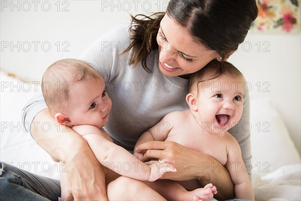 Caucasian mother holding twin baby daughters in lap on bed