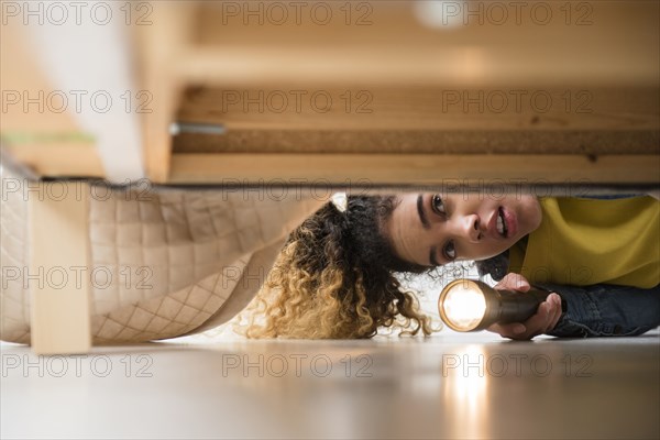 Mixed Race woman looking under bed with flashlight