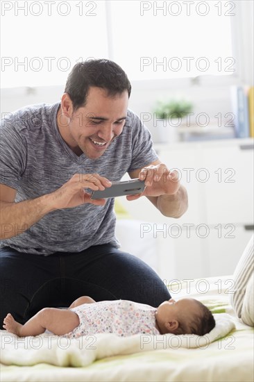 Hispanic father on bed photographing baby daughter with cell phone