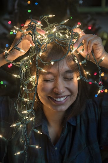 Mixed Race woman holding string lights on head near Christmas tree